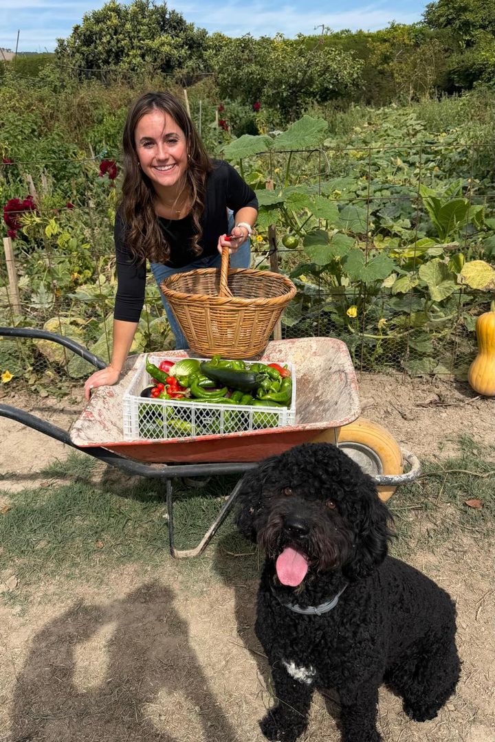 Cris en un huerto, sonriendo junto a una carretilla con pimientos y calabacines recién cogidos, una cesta de mimbre y su perro